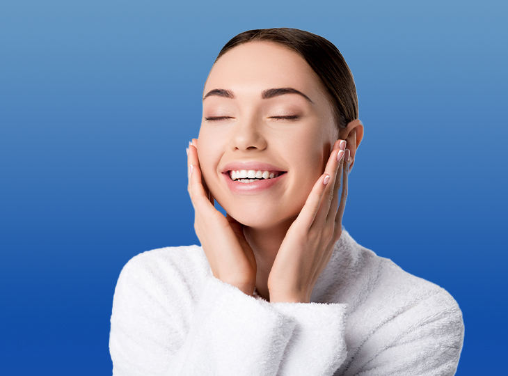 Woman in white sweater holding her face while smiling with eyes closed on blue background
